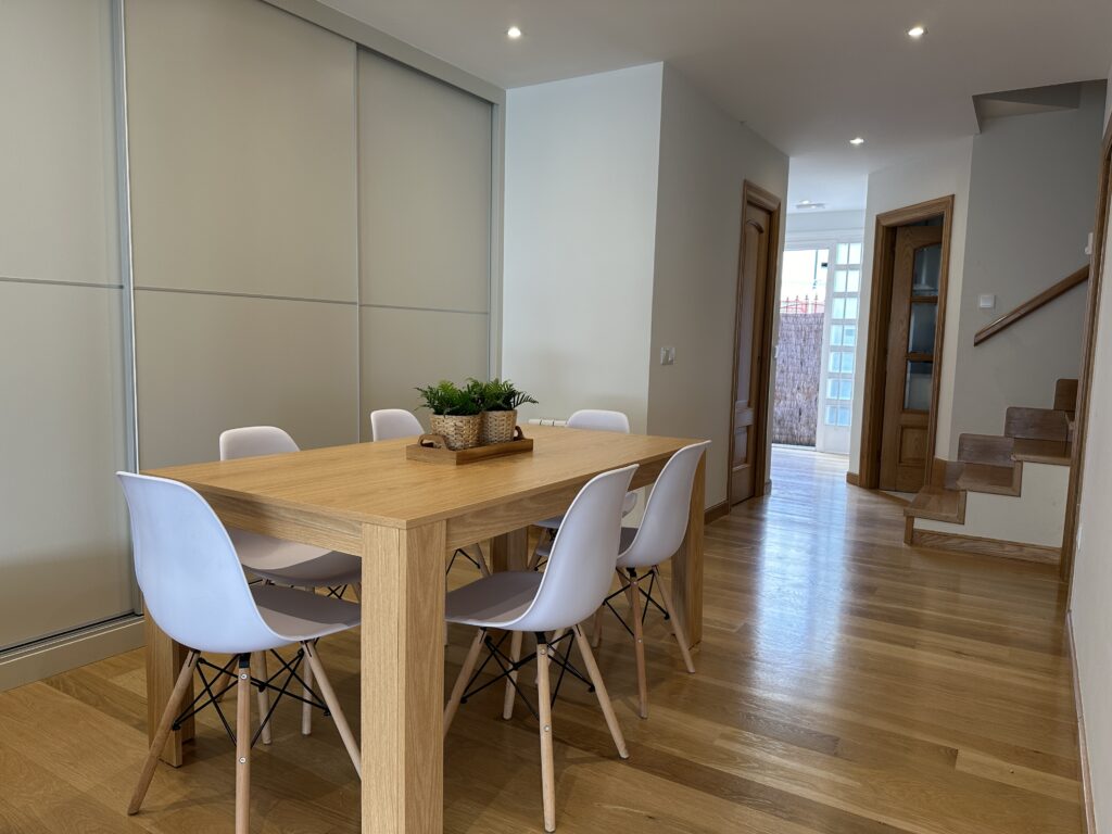 Comedor con mesa de madera, sillas blancas y pasillo con escalera al fondo